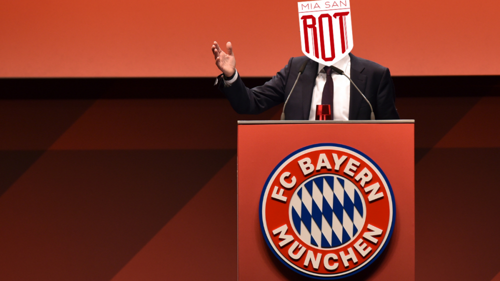 Präsident Herbert Hainer ist bei einer Jahreshauptversammlung hinter einem Podium zu sehen. Auf dem Podium ist ein Bayern-Logo abgebildet, er selbst trägt einen dunklen Anzug, mit hellem Hemd und dunkelroter Krawatte. Über seinen Kopf ist das Miasanrot-Logo rein retuschiert.