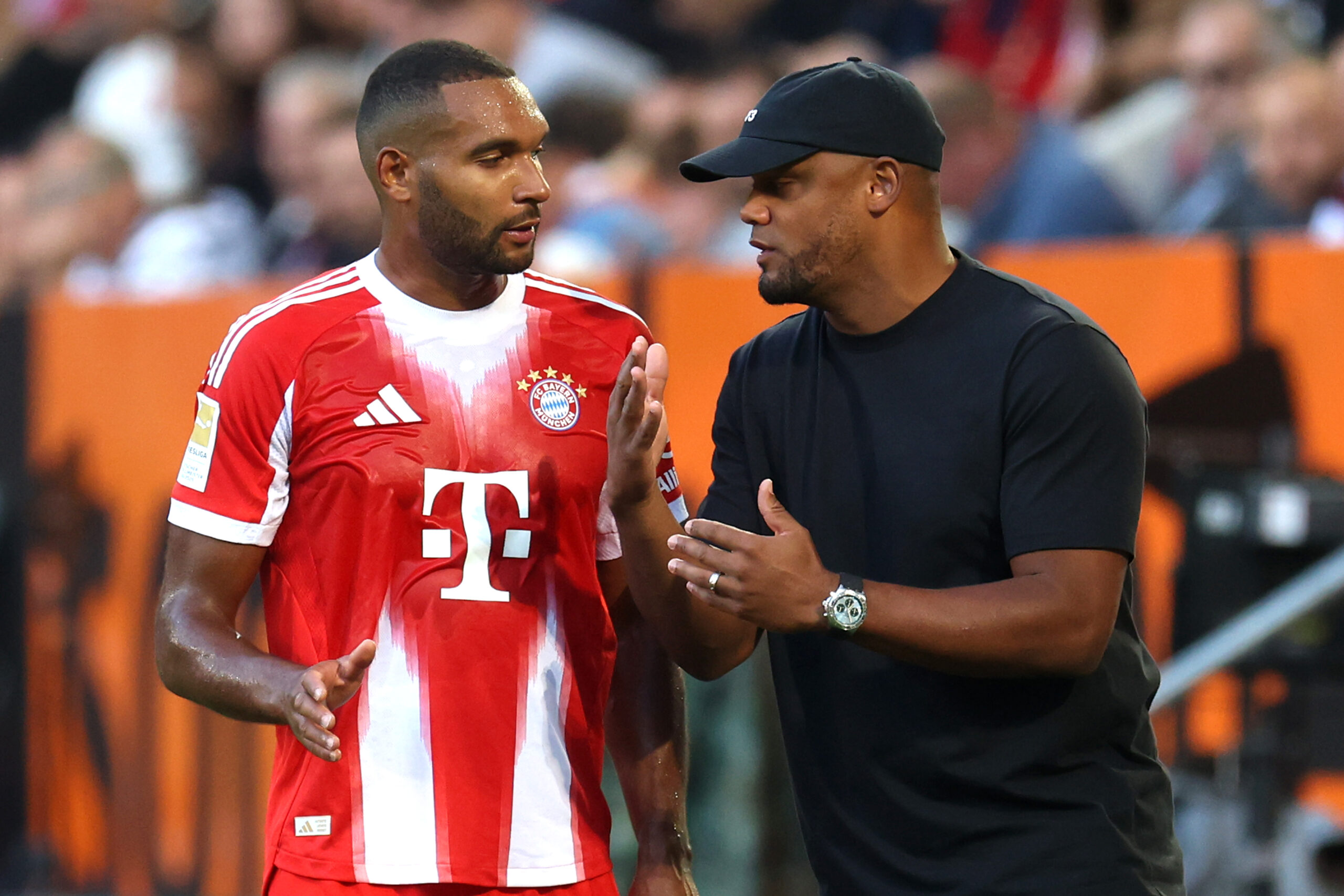 Vincent Kompany im Gespräch mit Jonathan Tah. Tah trägt das rot-weiße Trikot des FC Bayern, Kompany ist dunkel gekleidet.