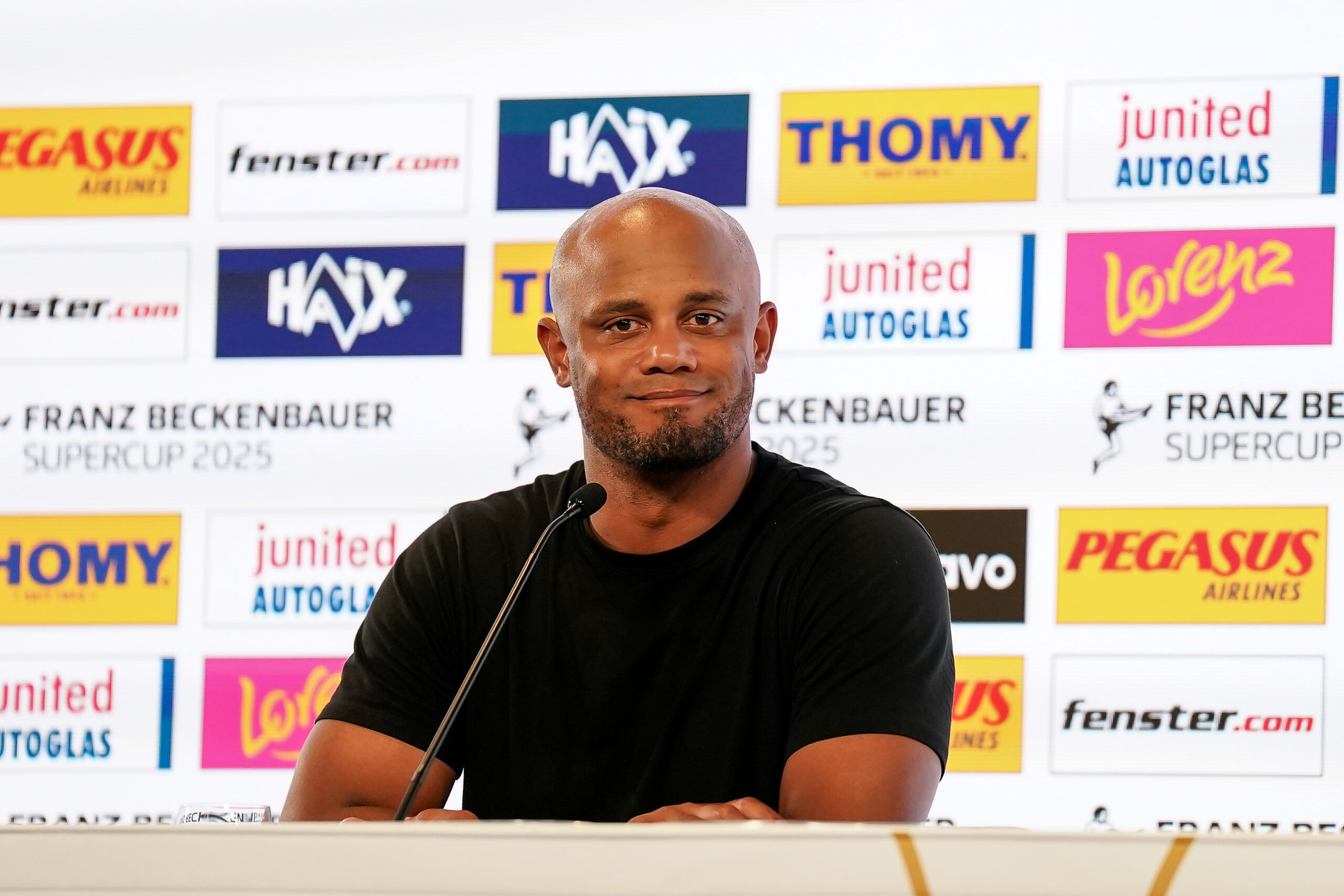 Vincent Kompany vom FC Bayern München sitzt grinsend auf der Pressekonferenz.