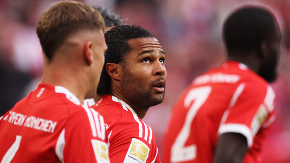 Serge Gnabry im roten Trikot des FC Bayern München. Er schaut nach oben, um ihn herum laufen Kimmich und Upamecano