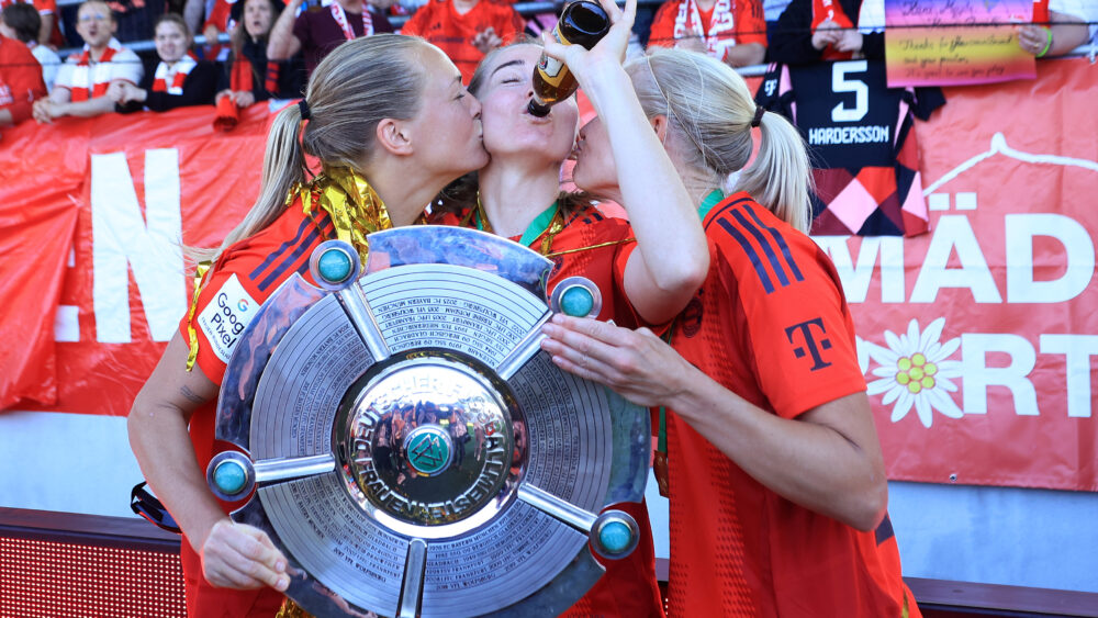 Magdalena Eriksson und Pernille Harder küssen Linda Sembrant auf die Wange. Sembrant trinkt aus einer Flasche. Die drei halten die Meisterschale in der Hand, die sie mit dem FC Bayern gewonnen haben. Im Hintergrund Fans.