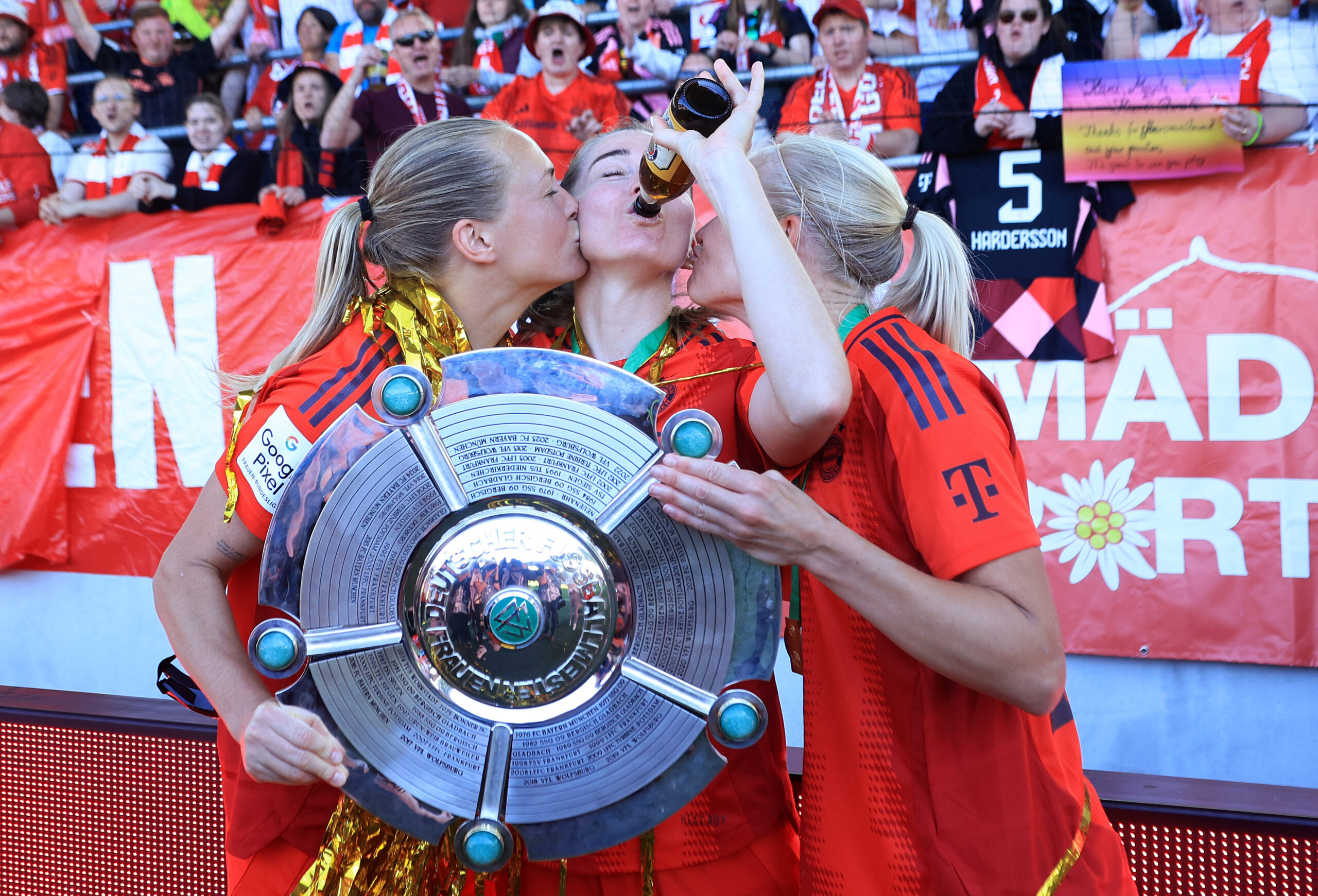 Magdalena Eriksson und Pernille Harder küssen Linda Sembrant auf die Wange. Sembrant trinkt aus einer Flasche. Die drei halten die Meisterschale in der Hand, die sie mit dem FC Bayern gewonnen haben. Im Hintergrund Fans.