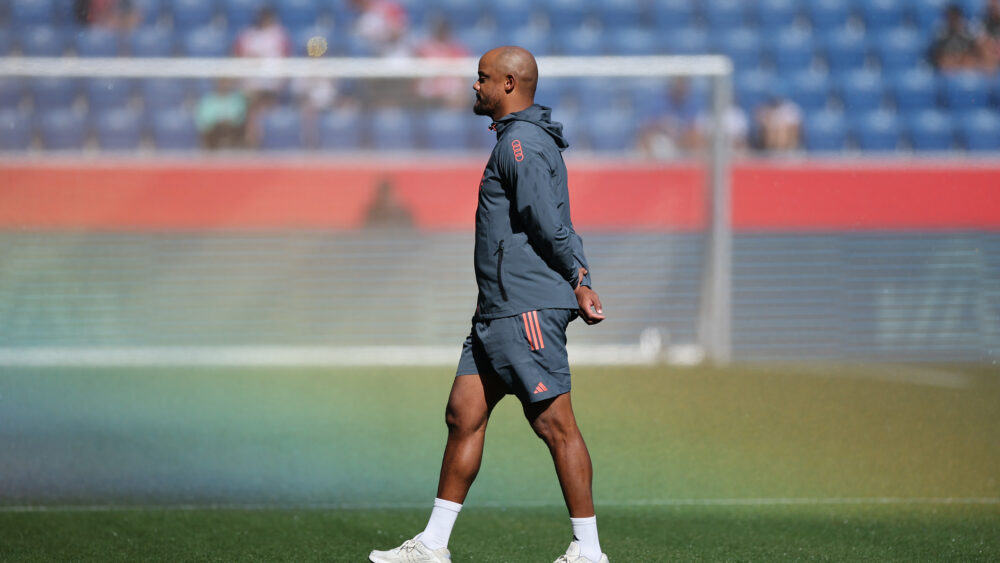 Vincent Kompany läuft über den Rasen. Der Trainer des FC Bayern hat die Arme hinter dem Rücken verschränkt.