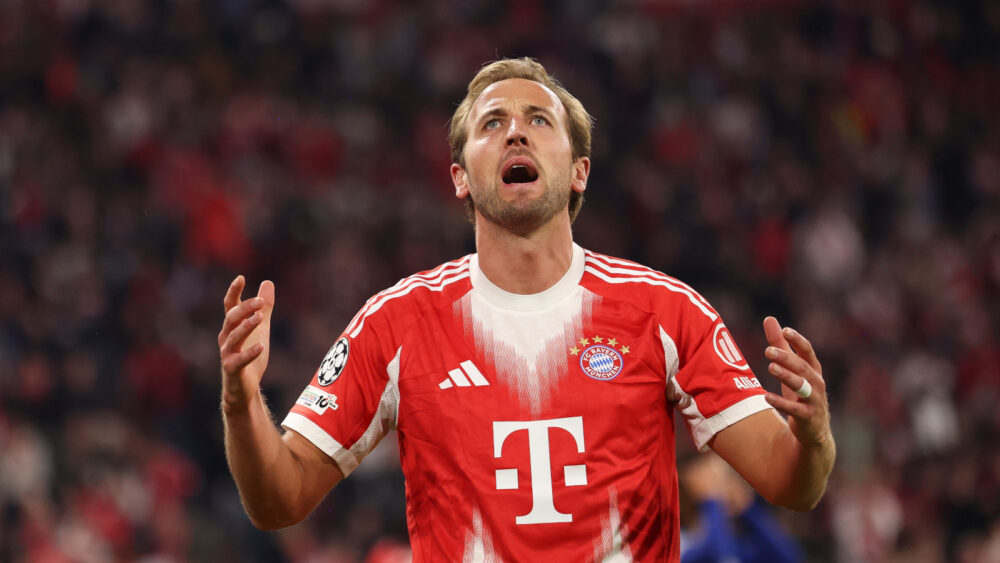 Harry Kane trägt ein rot-weißes Trikot des FC Bayern. Er hadert und hebt die Arme.