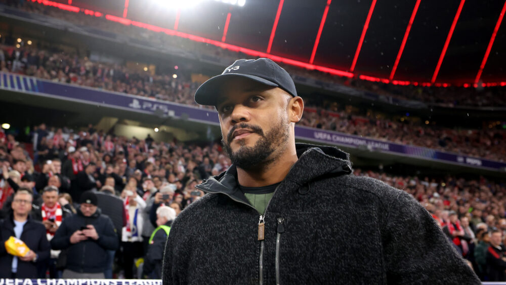 Vincent Kompany in der Allianz Arena des FC Bayern. Die Kamera filmt ihn von unten, er trägt dunkle Kleidung und eine dunkle Mütze. Hinter ihm das Publikum in der Arena.