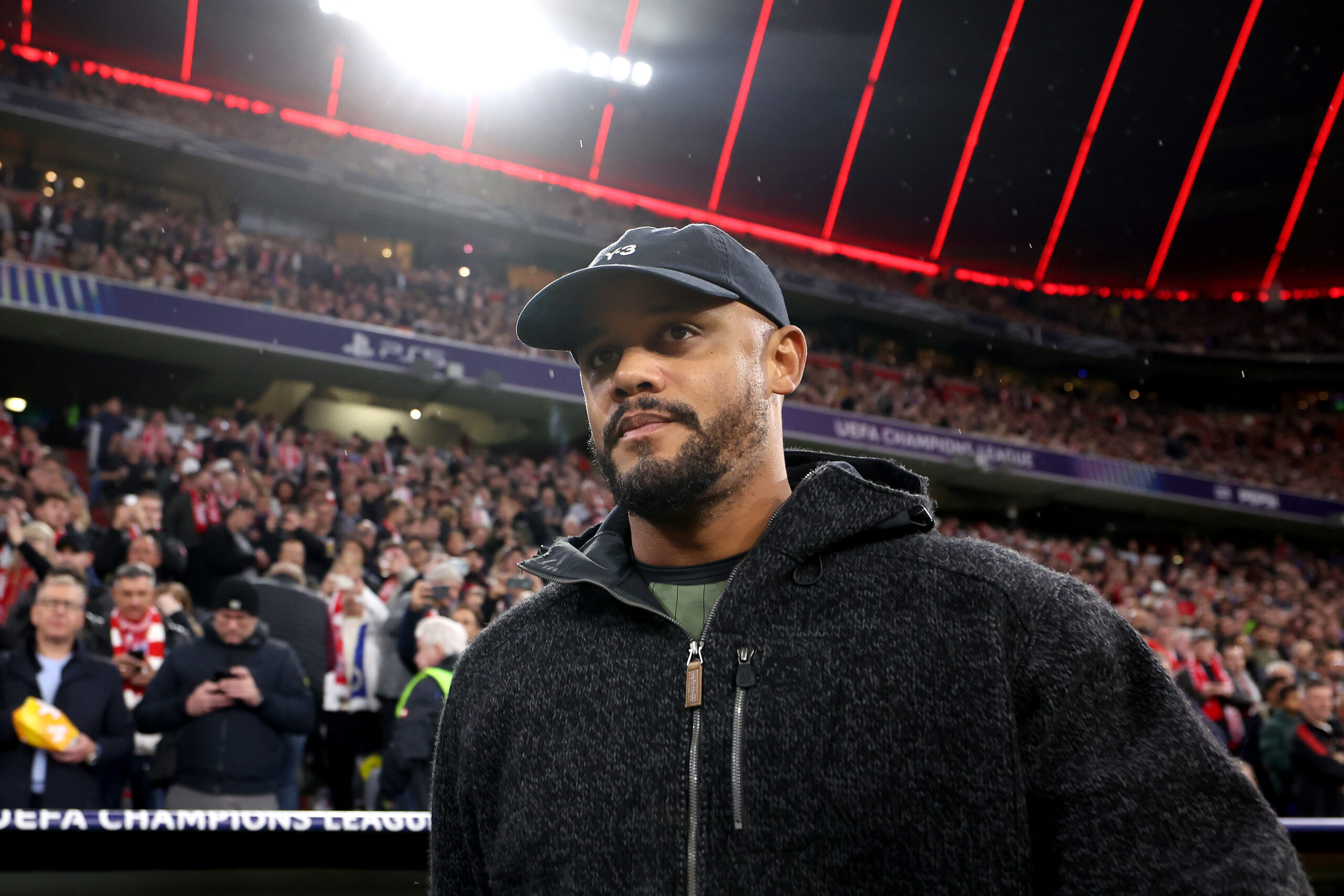 Vincent Kompany in der Allianz Arena des FC Bayern. Die Kamera filmt ihn von unten, er trägt dunkle Kleidung und eine dunkle Mütze. Hinter ihm das Publikum in der Arena.