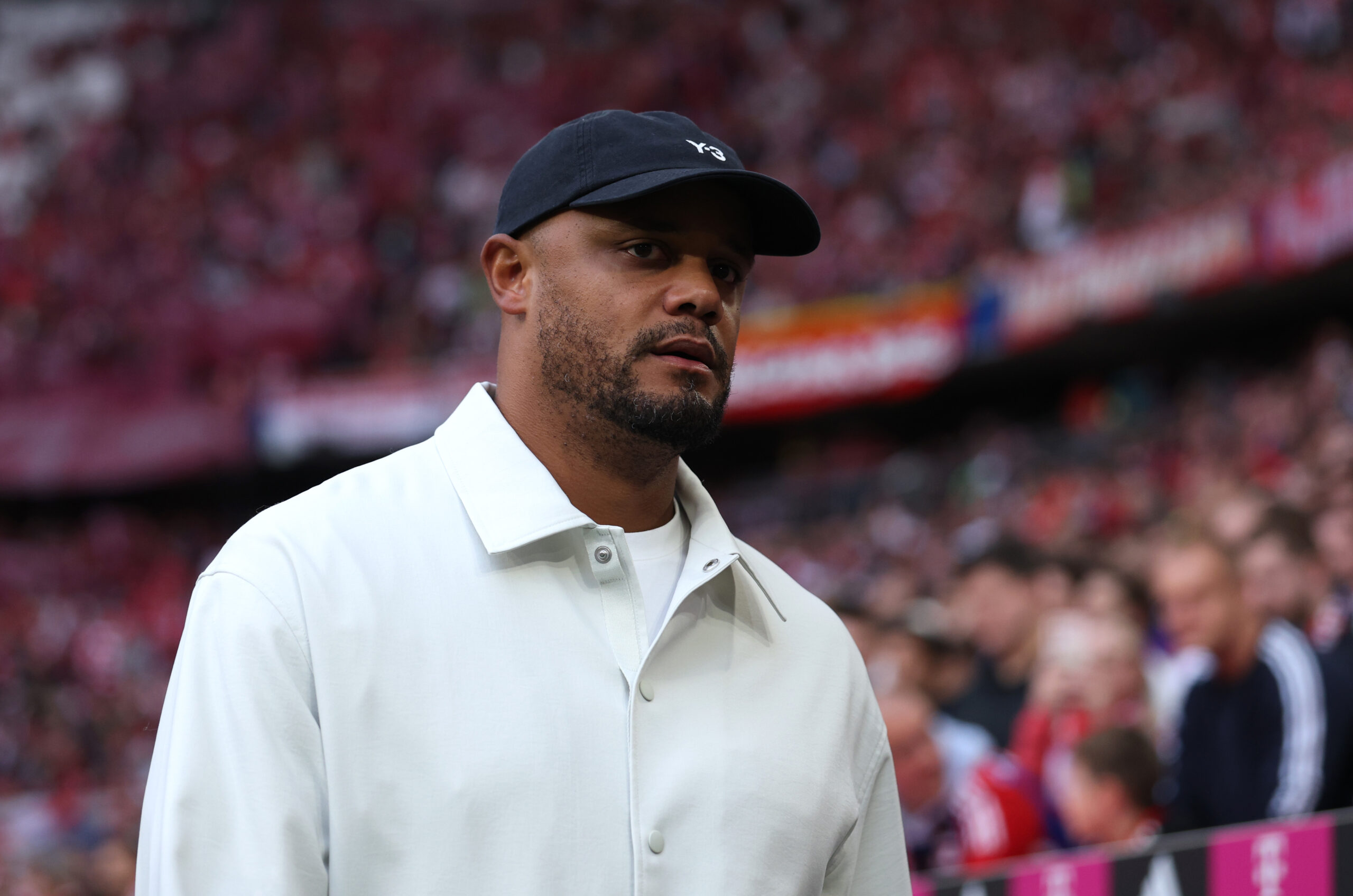 Vincent Kompany im Stadion des FC Bayern München. Er trägt helle Kleidung und eine dunkle Mütze.