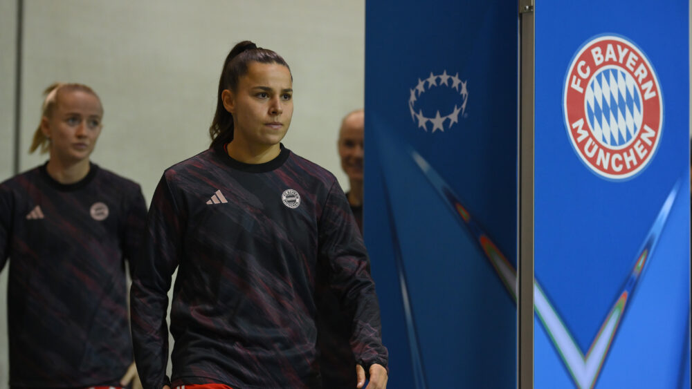 Lena Oberdorf und Lea Schüller laufen in Trainingsjacken durch den Innenraum. Rechts ist eine blaue Säule mit dem Logo der Champions League und dem Logo der FC Bayern Frauen bzw. des FC Bayern München.