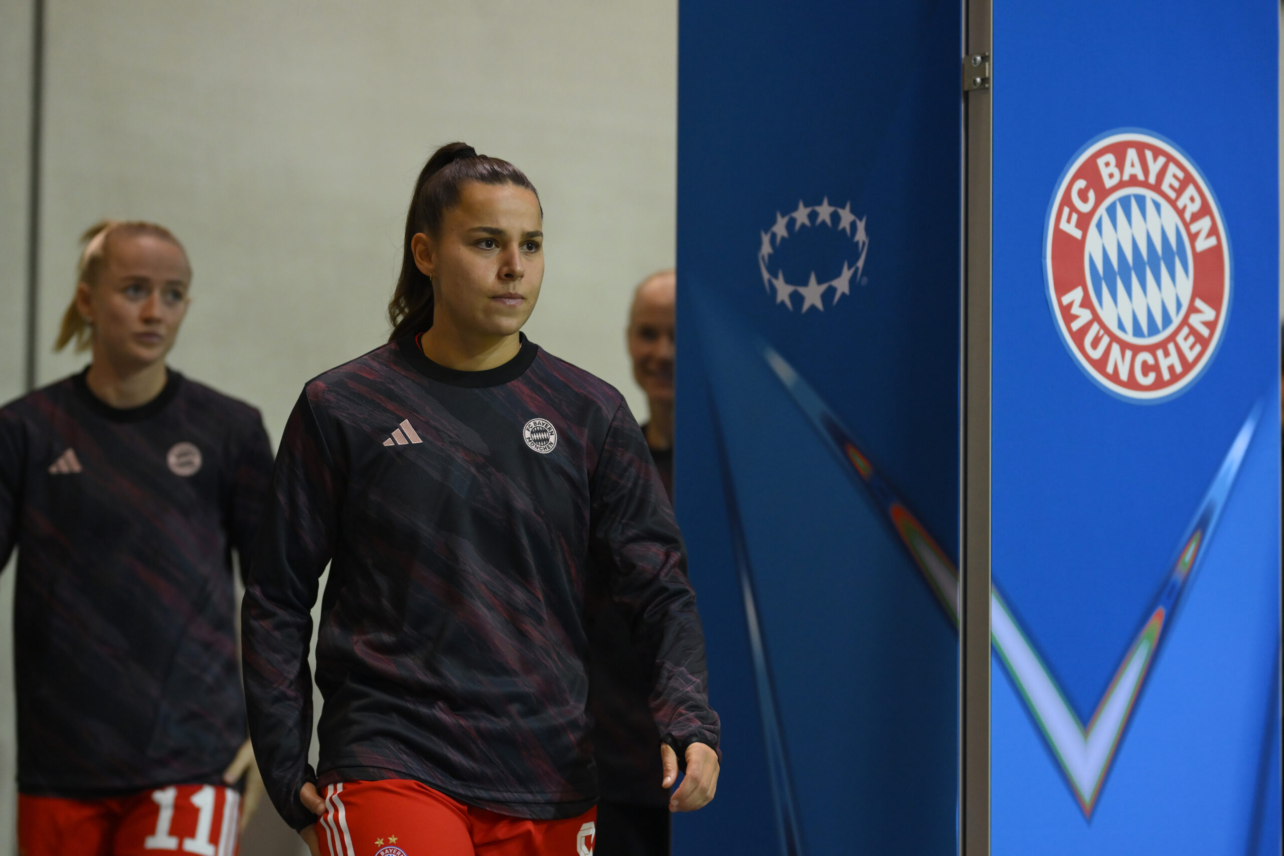 Lena Oberdorf und Lea Schüller laufen in Trainingsjacken durch den Innenraum. Rechts ist eine blaue Säule mit dem Logo der Champions League und dem Logo der FC Bayern Frauen bzw. des FC Bayern München.