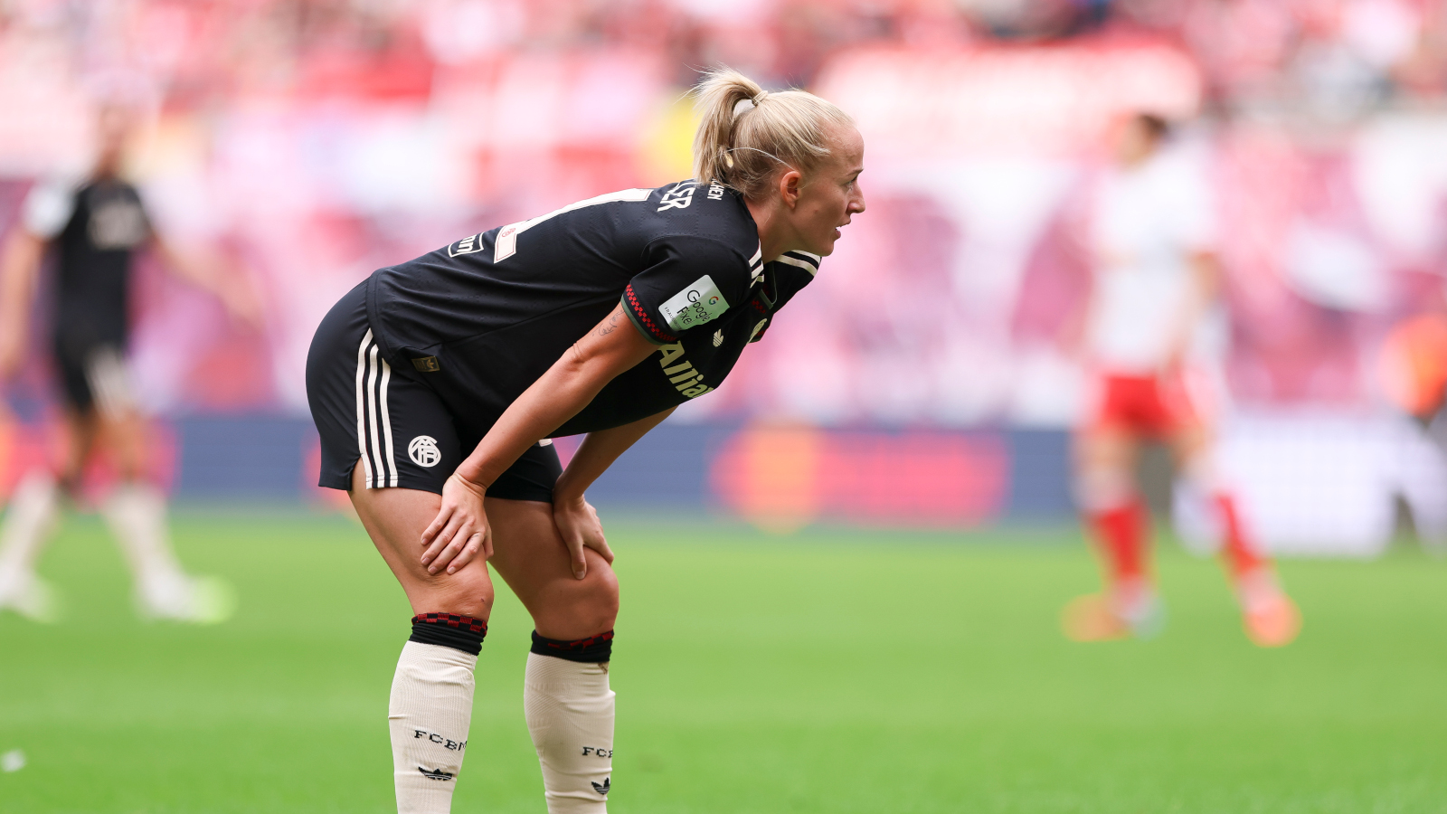 Lea Schüller im dunklen Trikot des FC Bayern. Sie bückt sich und stützt sich auf ihre Knie.