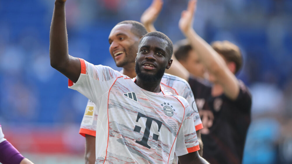Dayot Upamecano hebt den linken Arm, hinter ihm steht sein Kollege vom FC Bayern München Jonathan Tah und lacht