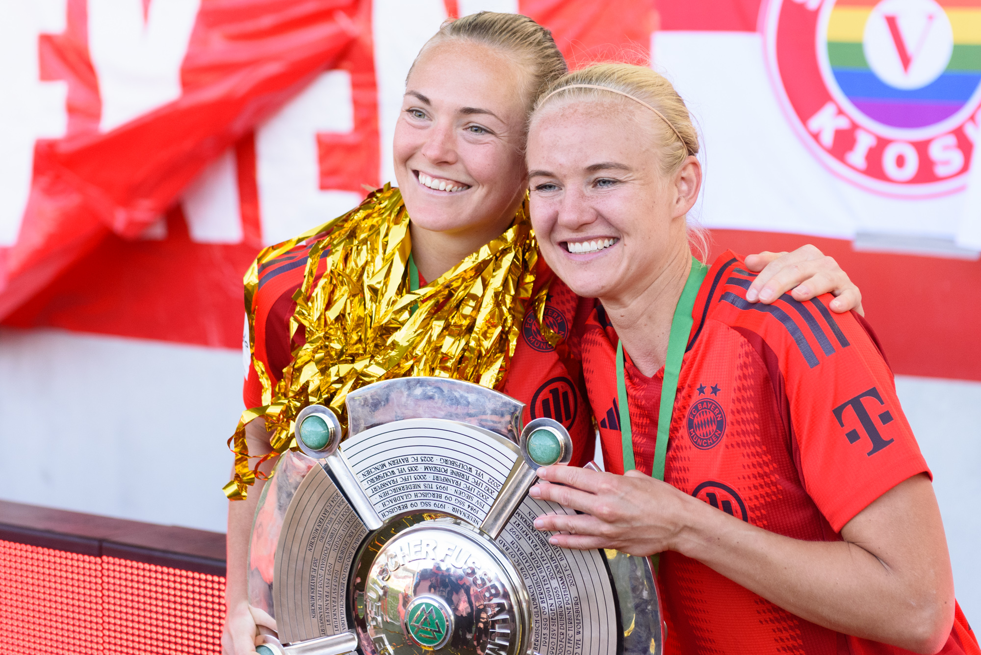 Magdalena Eriksson und Pernille Harder Arm in Arm mit der Meisterschale. Eriksson hat viel goldenes Konfetti um den Hals, beide lächeln und tragen das rote Heimtrikot der FC Bayern Frauen der Saison 2024/25.