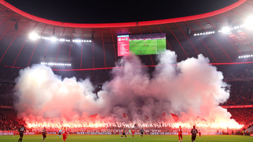 Die Südkurve der Allianz Arena ist im Hintergrund zu sehen. Von dort steigt Rauch auf, der von den Feuern der Bengalos kommt, die die Fans des FC Bayern gezündet haben. Davor findet das Spiel zwischen dem FCB und Sporting statt.