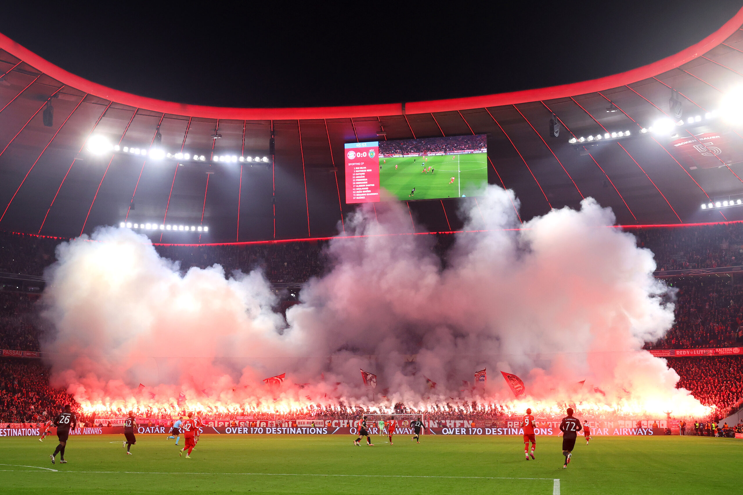 Die Südkurve der Allianz Arena ist im Hintergrund zu sehen. Von dort steigt Rauch auf, der von den Feuern der Bengalos kommt, die die Fans des FC Bayern gezündet haben. Davor findet das Spiel zwischen dem FCB und Sporting statt.
