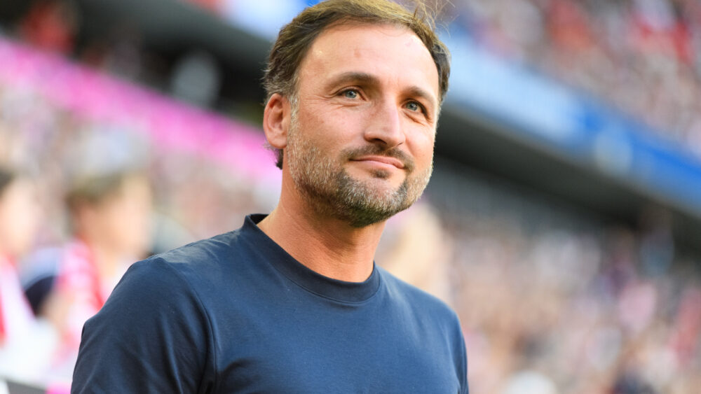 José Barcala (FC Bayern Frauen) steht grinsend an der Seitenlinie. Hinter ihm das Publikum in der Allianz Arena.
