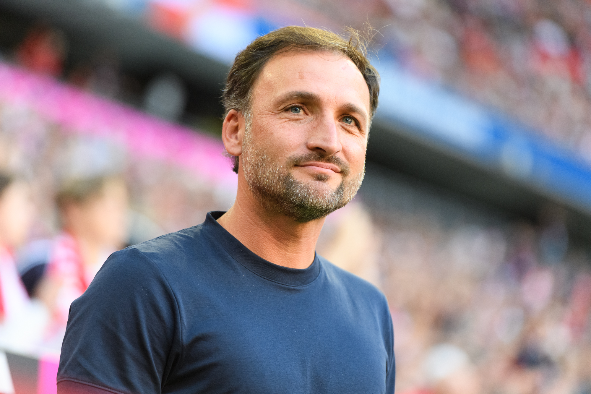 José Barcala (FC Bayern Frauen) steht grinsend an der Seitenlinie. Hinter ihm das Publikum in der Allianz Arena.