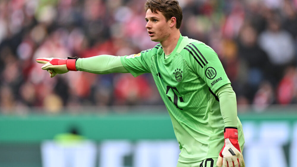 Jonas Urbig im grünen Torwarttrikot des FC Bayern: Er streckt den rechten Arm nach vorn und zeigt mit dem Zeigefinger in diese Richtung.