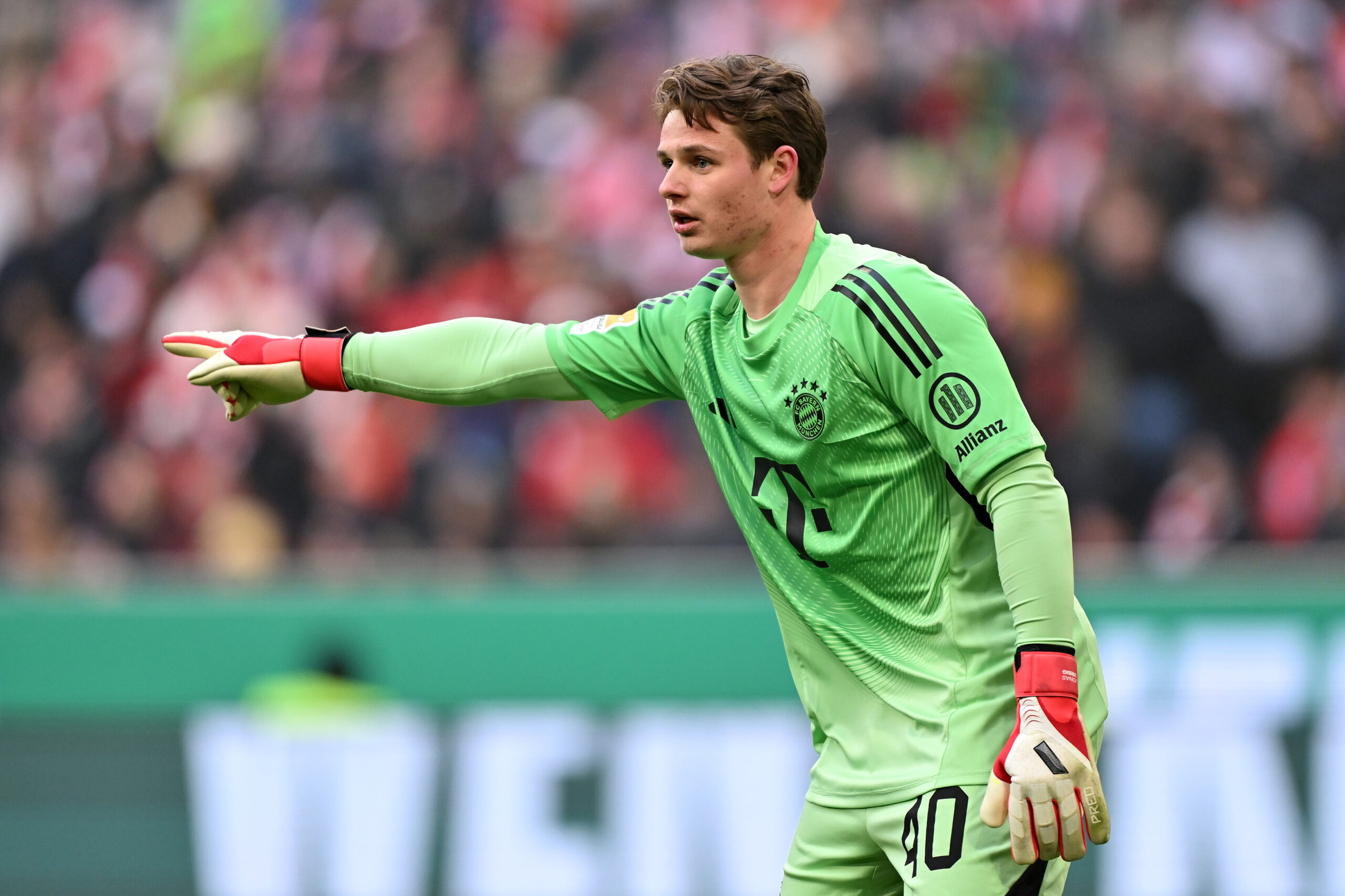 Jonas Urbig im grünen Torwarttrikot des FC Bayern: Er streckt den rechten Arm nach vorn und zeigt mit dem Zeigefinger in diese Richtung.