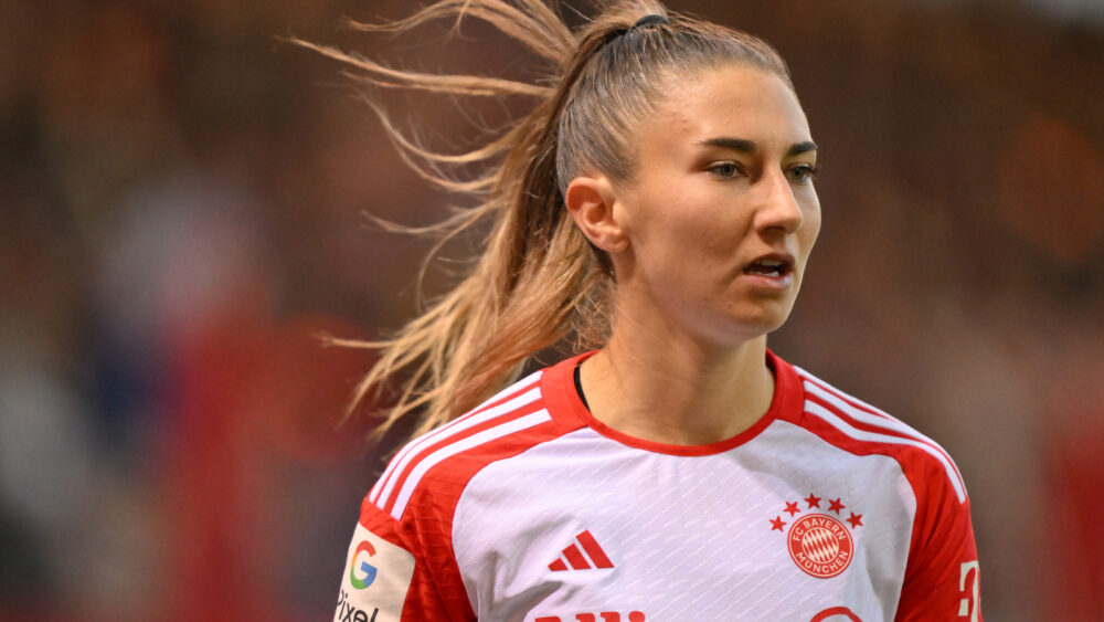 Katharina Naschenweng im rot-weißen Trikot der FC Bayern Frauen aus der Saison 2024/25, ihre Haare flattern im Wind, sie schaut fokussiert nach vorn.