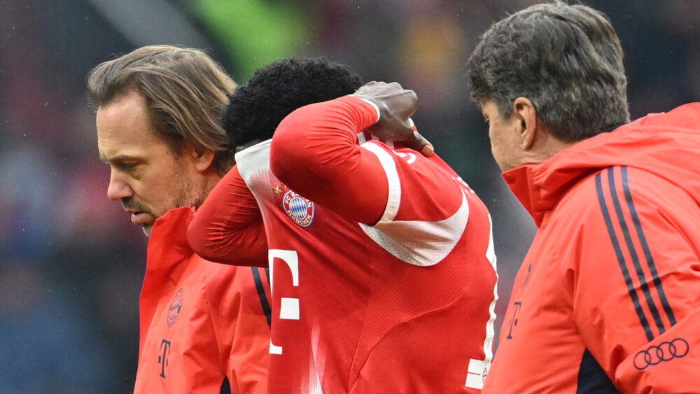 Alphonso Davies (FC Bayern) zieht sich das Trikot über den Kopf und wird von zwei Staffmitgliedern des FCB begleitet.