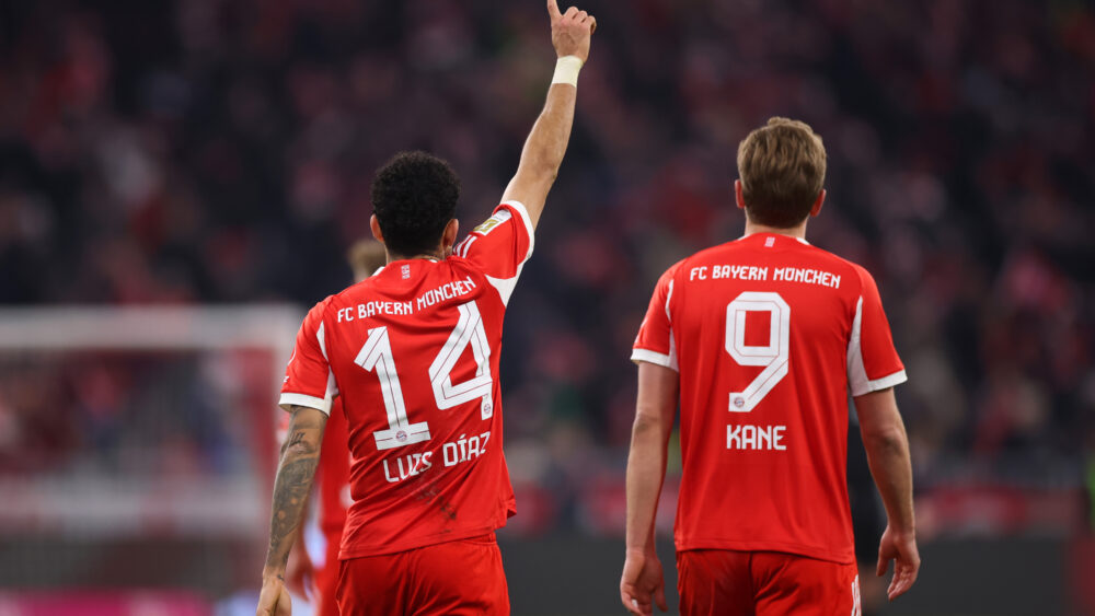 Luis Díaz und Harry Kane vom FC Bayern sind von hinten zu sehen. Díaz hebt den rechten Finger in die Luft.