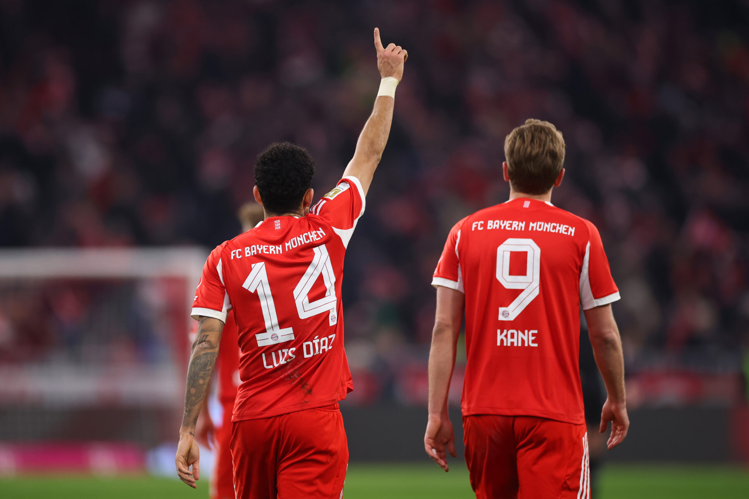 Luis Díaz und Harry Kane vom FC Bayern sind von hinten zu sehen. Díaz hebt den rechten Finger in die Luft.