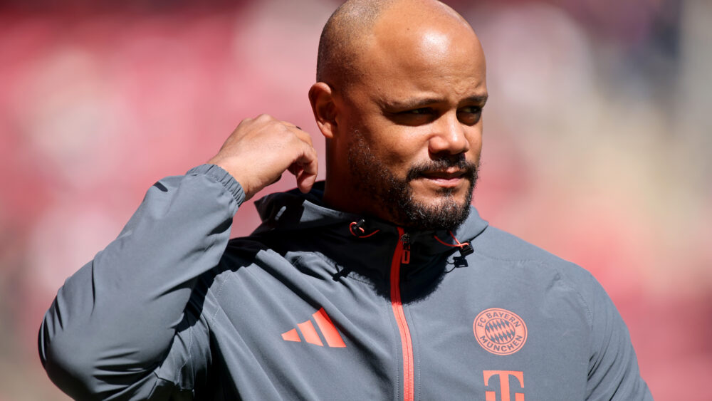 Vincent Kompany steht im grauen Trainingsanzug des FC Bayern in der Sonne und fasst sich mit einer Hand in Richtung Ohr.