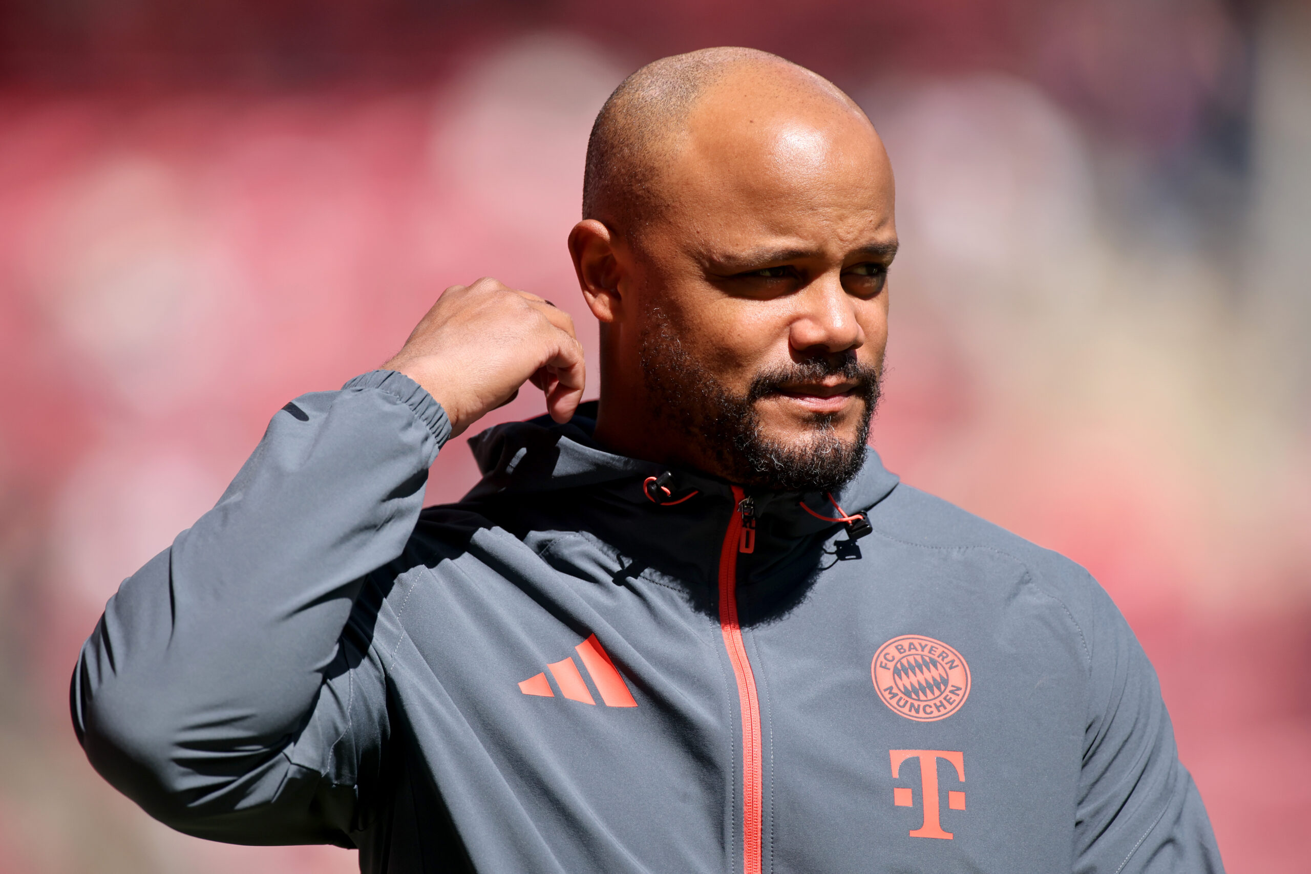Vincent Kompany steht im grauen Trainingsanzug des FC Bayern in der Sonne und fasst sich mit einer Hand in Richtung Ohr.