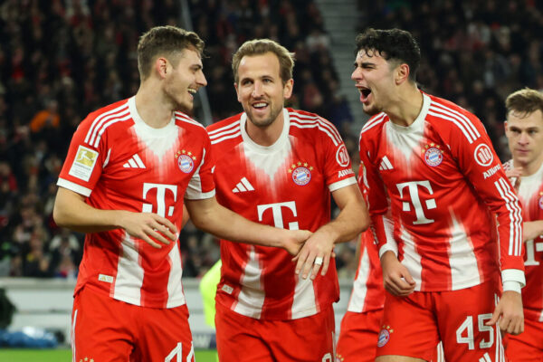 Josip Stanisic, Harry Kane und Aleksandar Pavlovic jubeln im Trikot des FC Bayern. Stanisic schaut auf die beiden anderen und lacht, Kane ist in der Mitte und lacht, Pavlovic scheint etwas laut zu rufen.