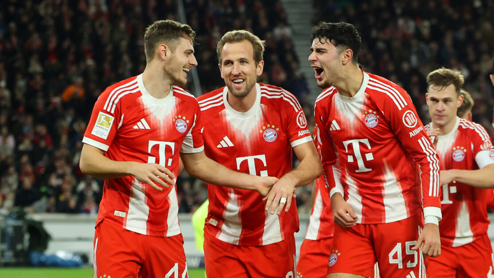Josip Stanisic, Harry Kane und Aleksandar Pavlovic jubeln im Trikot des FC Bayern. Stanisic schaut auf die beiden anderen und lacht, Kane ist in der Mitte und lacht, Pavlovic scheint etwas laut zu rufen.