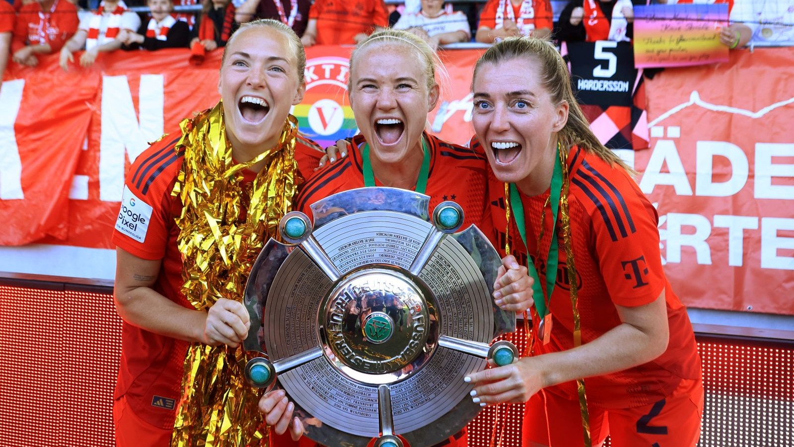 Magdalena Eriksson, Pernille Harder und Linda Sembrant mit der Meisterschale und goldenem Konfetti. Sie tragen die Trikots des FC Bayern, hinter ihnen sind Banner von Fans zu sehen.