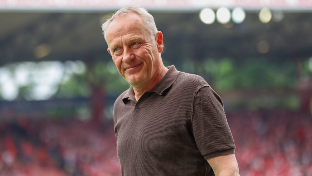 Christian Streich (Experte bei der WM 2026) vor einer Tribüne. Er grinst leicht.