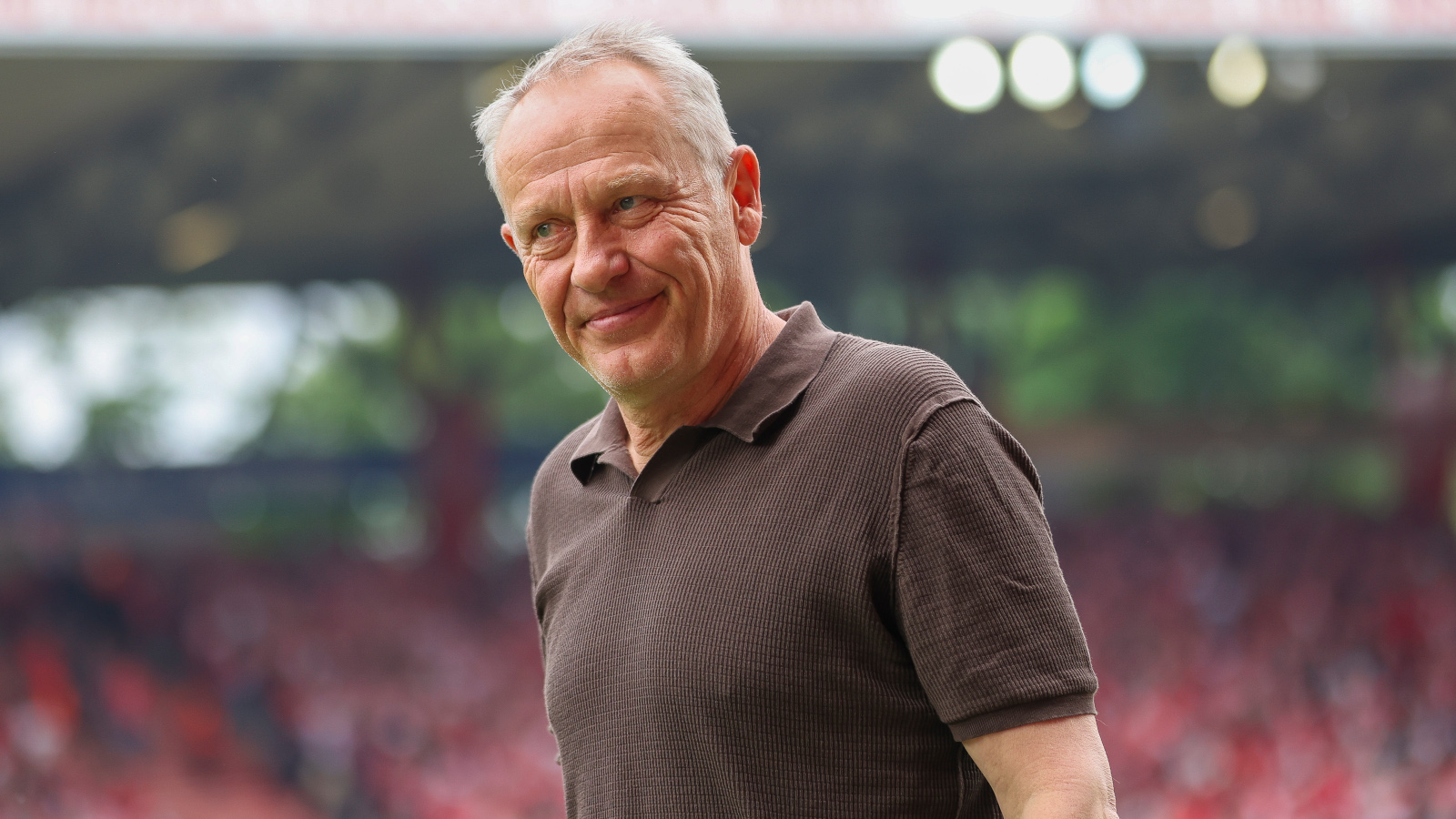Christian Streich (Experte bei der WM 2026) vor einer Tribüne. Er grinst leicht.