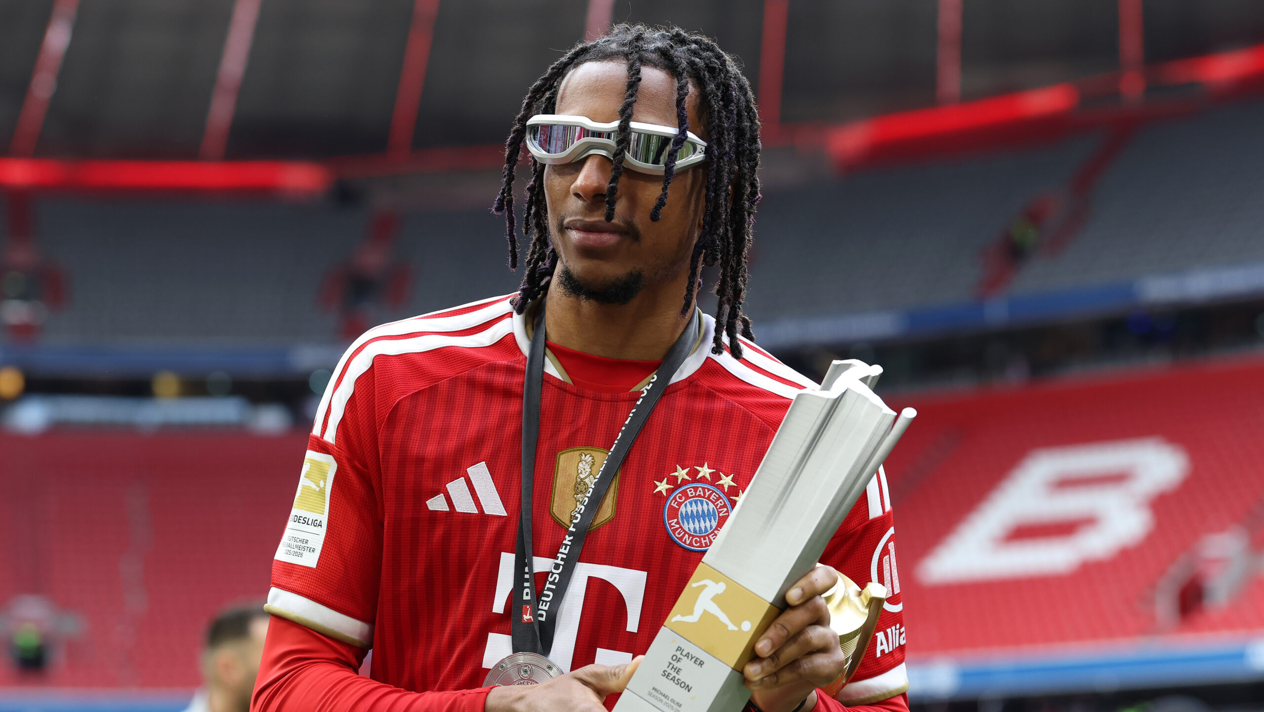Michael Olise (FC Bayern) mit einer individuellen Auszeichnung in der Hand und einer schnellen Brille im Gesicht.