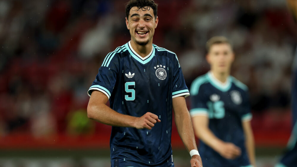Aleksandar Pavlovic (FC Bayern) im blauen Deutschland-Trikot. Er zwinkert und lacht.