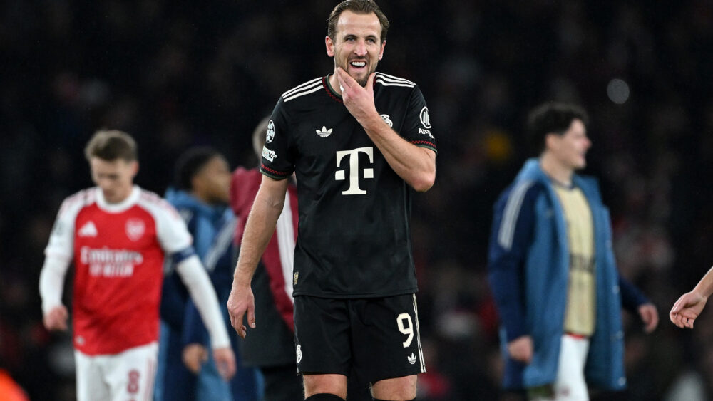 Harry Kane greift sich ins Gesicht und verzieht das Gesicht. Er trägt ein schwarzes trikot des FC Bayern. Im Hintergrund sind Spieler des FC Arsenal zu sehen.