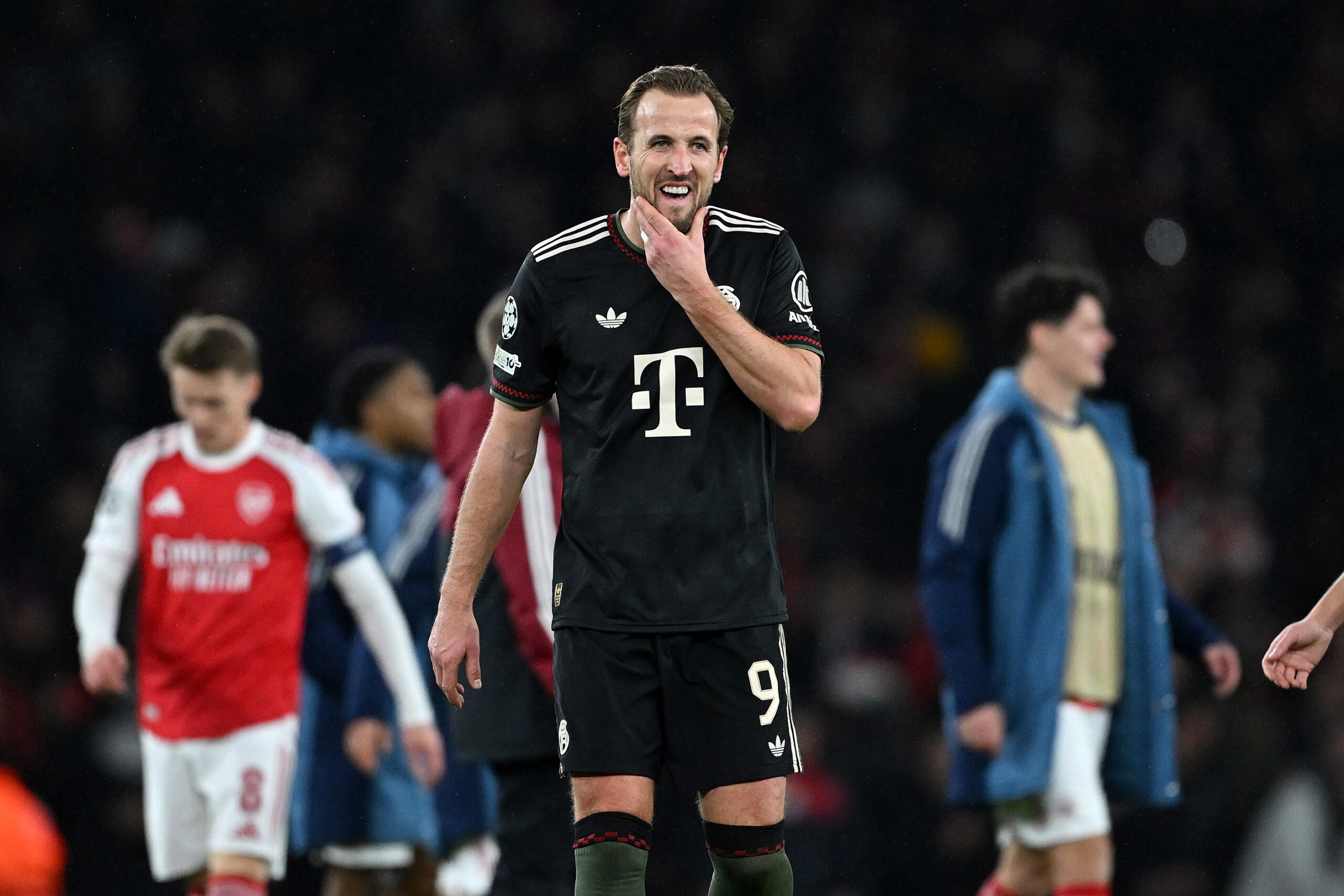 Harry Kane greift sich ins Gesicht und verzieht das Gesicht. Er trägt ein schwarzes trikot des FC Bayern. Im Hintergrund sind Spieler des FC Arsenal zu sehen.